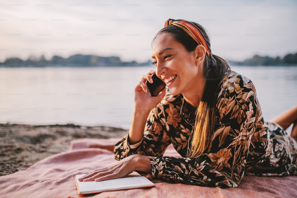 Attractive caucasian brunette in floral dress and with headband on head lying on stomach on picnic blanket on the shore and talking on the cellphone.
