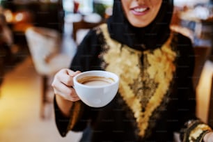 Close up de mulher árabe sorridente vestida em trajes tradicionais sentada no café e segurando uma xícara de café fresco. Tempo livre nos fins de semana.
