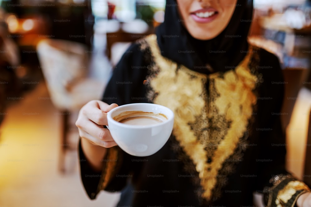 전통 의상을 입고 카페에 앉아 신선한 에스프레소 한 잔을 들고 웃고 있는 아랍 여성의 클로즈업. 주말에는 자유 시간.