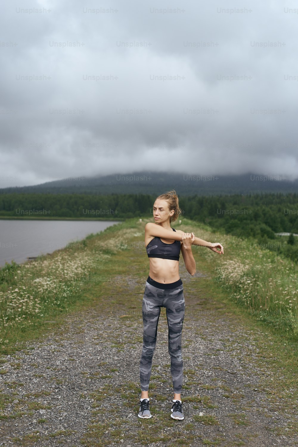 Mid age skinny sportswoman exercising outdoor in summer, on gloomy day, standing at the road with scenic view, doing warm up stretching, wearing tank