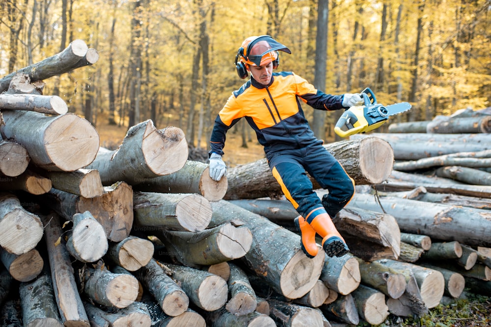 Professioneller Holzfäller in Arbeitsschutzkleidung springt mit einer Kettensäge von einem Baumstammhaufen im Wald
