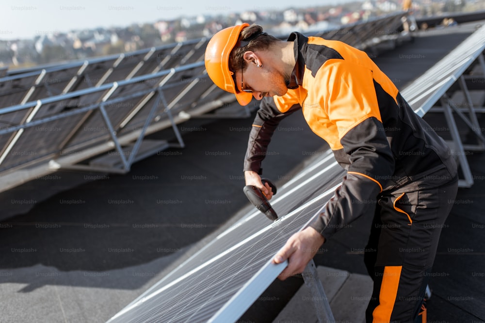 太陽光発電の屋上プラントにソーラーパネルを設置または交換するオレンジ色の保護服を着た設備の整った労働者。太陽光発電所の保守・設置コンセプト