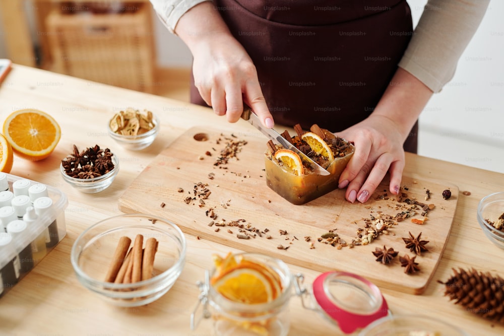 Hände der jungen Frau mit Messer schneiden große handgemachte Seifenstück mit aromatischen Gewürzen und Orangenscheiben auf Holzbrett