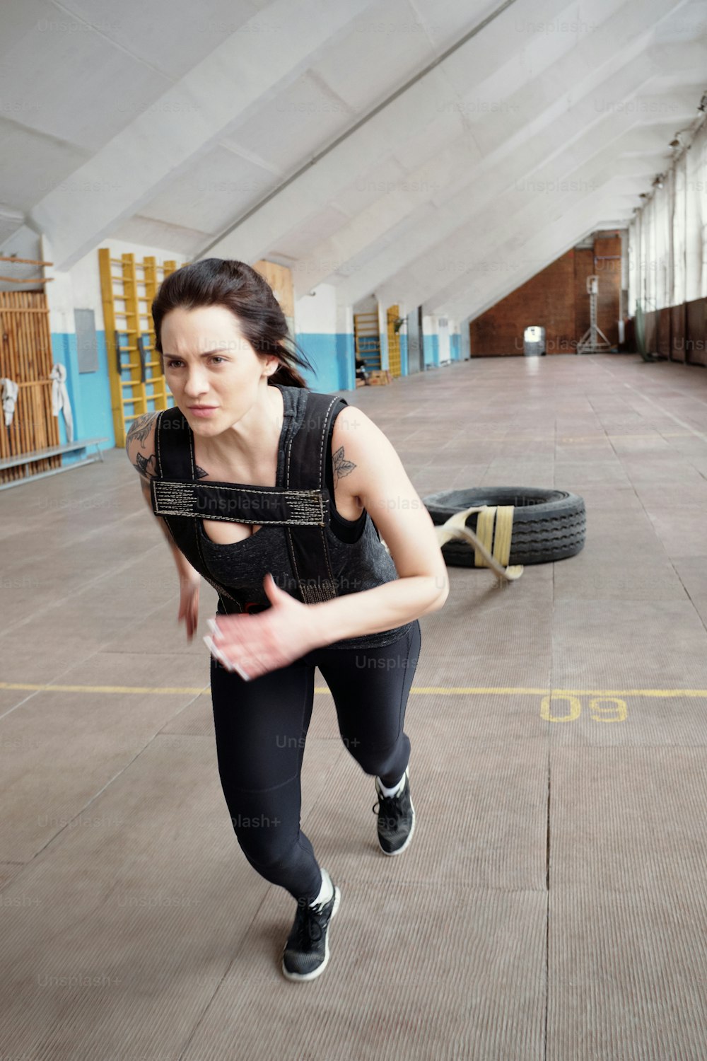 Tire pulling workout. Young attractive Caucasian sportswoman dragging tire with ropes while having cross training in sports hall