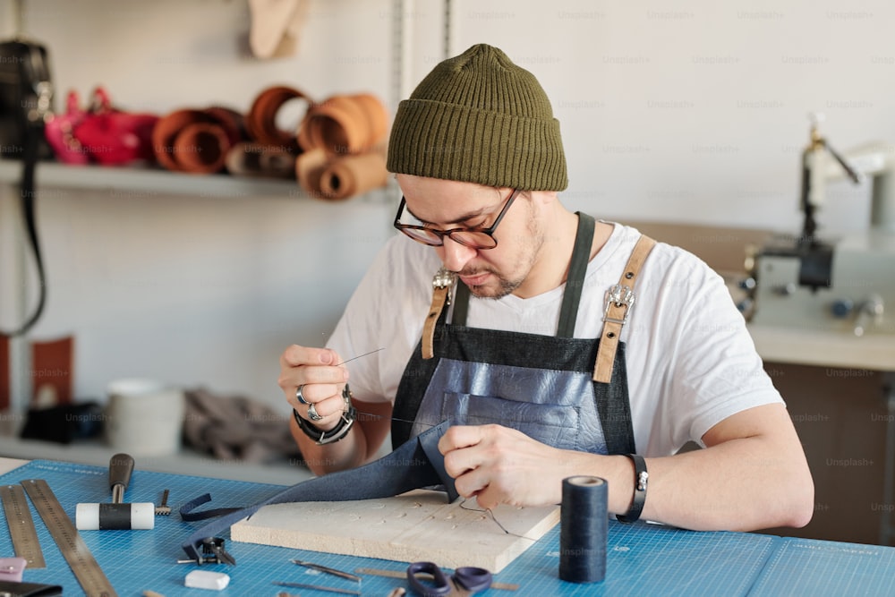 Jeune homme sérieux et beau utilisant une longue aiguille tout en cousant du cuir à la main dans un atelier