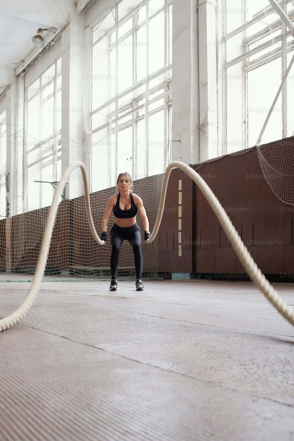 Battle rope workout. Young slim Caucasian woman in black sportswear burning calories by doing exercise with ropes in gym