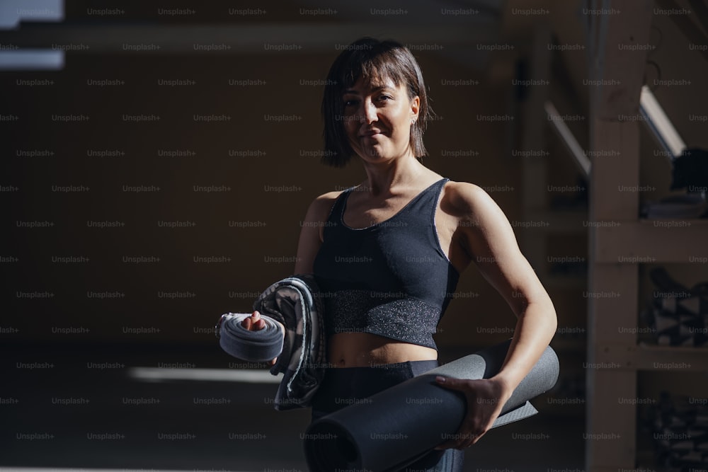 Fit slim smiling sportswoman standing in fitness studio, holding mat and fitness equipment.