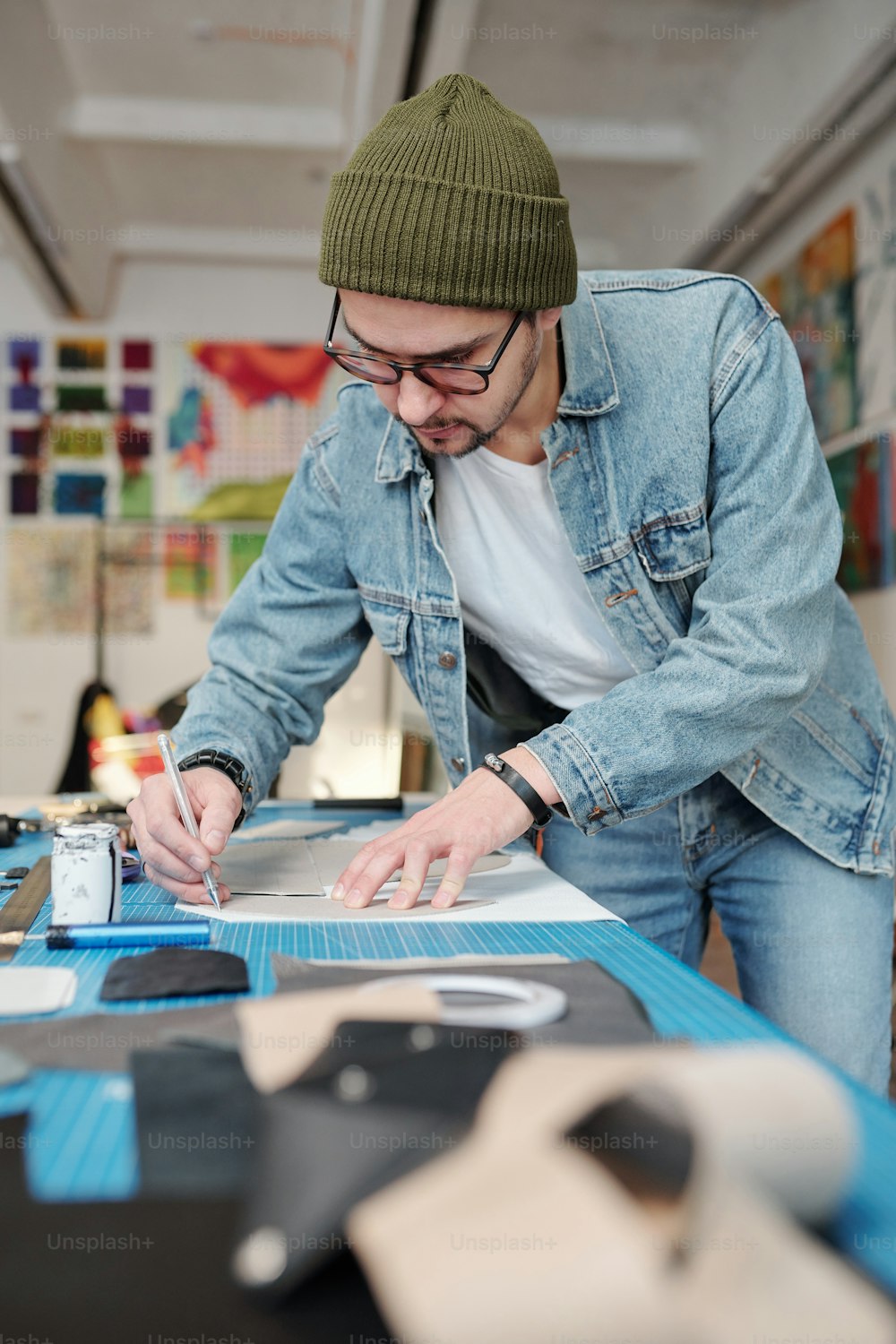 Fokussierter junger Mann im Denim-Outfit, der in der Werkstatt Nähmuster auf Lederstoff nachzeichnet