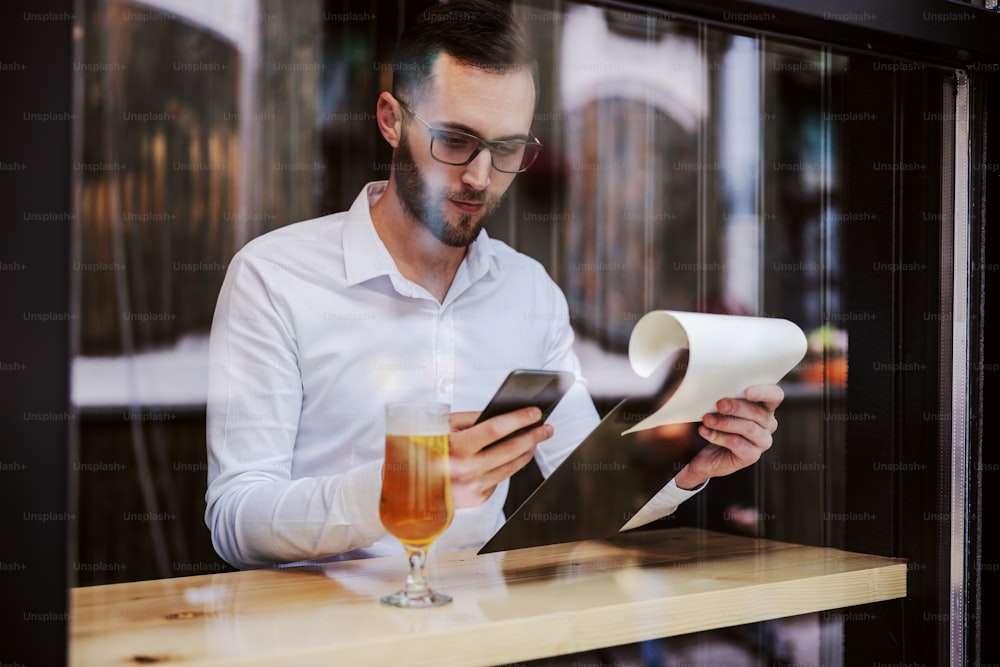 Young serious businessman in shirt sitting in pub after work, using smart phone for reading an e-mail and holding paperwork. Picture taken from outside.