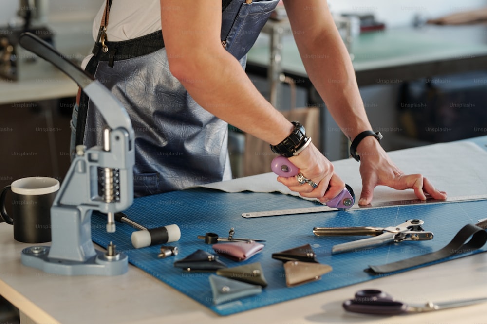 Close-up de homem irreconhecível em avental usando régua de metal para cortar borda de couro em estúdio de artesanato