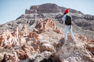 Young woman dressed casually in jeans with red hat and backpack hiking on the rocky terrain on a sunny day. Traveling on volcanic valley on Tenerife island