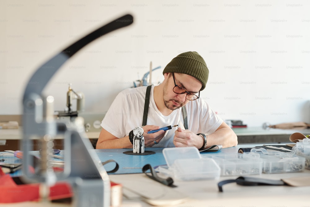 Serious young leather worker in hipster hat applying oil-dye on leather edge in workshop