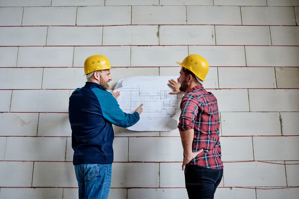 보호 헬멧을 쓴 두 명의 성공적인 건축업자가 스타트업 회의에서 미완성 건물의 벽 옆에 서서 청사진을 논의하고 있습니다.