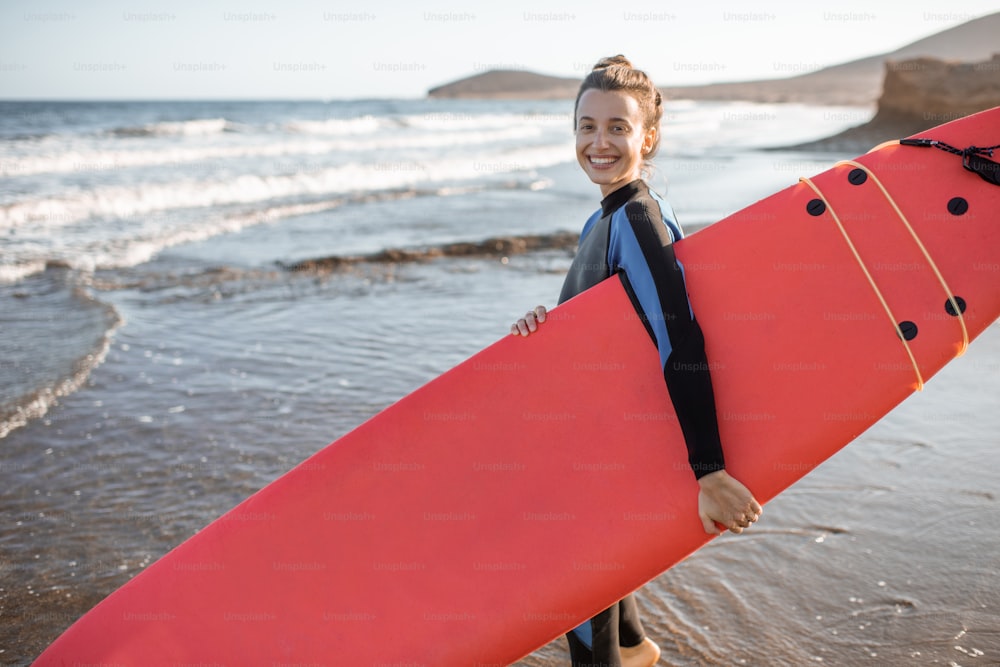 Portrait of a young woman in wetsuit standing with surfboard on the beautiful wild beach on a sunset. Water sport and active lifestyle concept