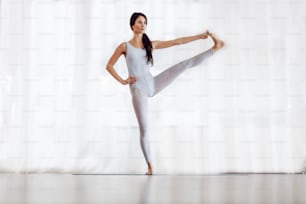 Full length of fit slim attractive young brunette with long hair standing barefoot in Extended Standing Hand to Toe yoga position. Yoga studio interior.