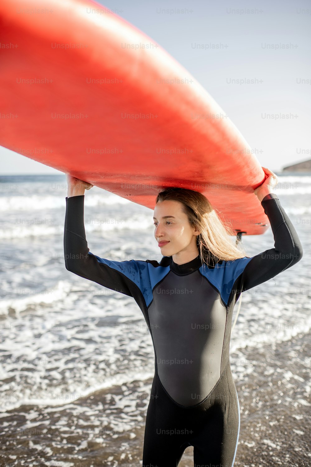 Porträt einer jungen Frau im Neoprenanzug, die Surboard über dem Kopf trägt und sich auf das Surfen am wilden Strand bei Sonnenuntergang vorbereitet. Aktives Lifestyle-Konzept