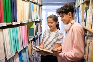 College student with tablet and his classmate looking through book in library while standing between bookshelves