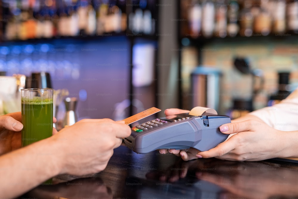 Hand of young man holding plastic card over payment machine held by waitress while paying for glass of fresh vegetable smoothie