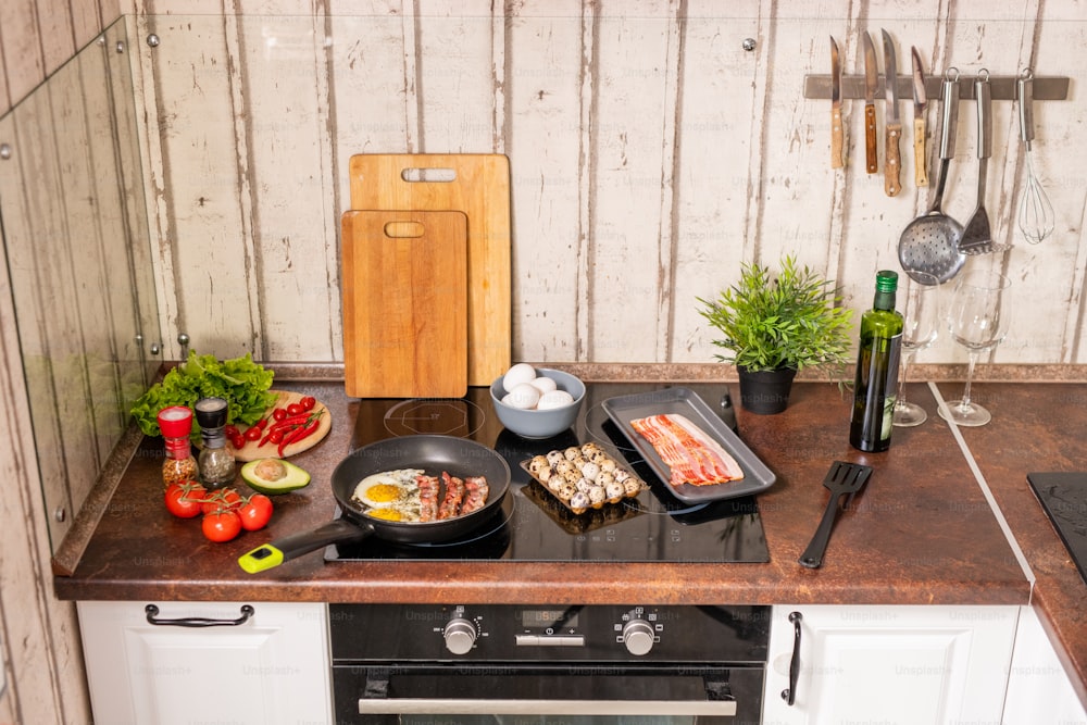 Electric stove with frying pan during cooking eggs with bacon surrounded by fresh fruits and vegetables, spices, wooden board and bottle of olive oil