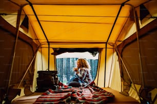 Une femme caucasienne adulte heureuse et solitaire s’assoit à l’extérieur d’une tente en profitant d’une tasse de thé et de la nature sauvage en plein air autour - vacances de camping alternatives gratuites avec tente et sac à dos pour les personnes dépendantes de l’envie de voyager