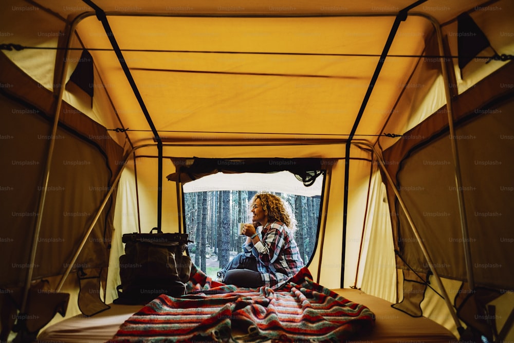 Glückliche einsame erwachsene kaukasische Frau setzt sich vor ein Zelt und genießt eine Tasse Tee und die wilde Natur im Freien - kostenloser alternativer Campingurlaub mit Zelt und Rucksack für Fernwehsüchtige