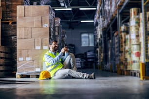 Young smiling employee sitting on the floor in warehouse, taking a break and using smart phone.