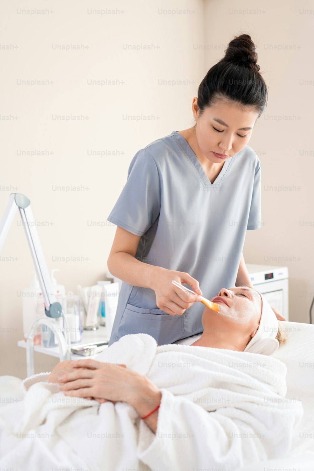 Asian beautician in scrubs applying facial mask on mature clients face using brush in beauty salon