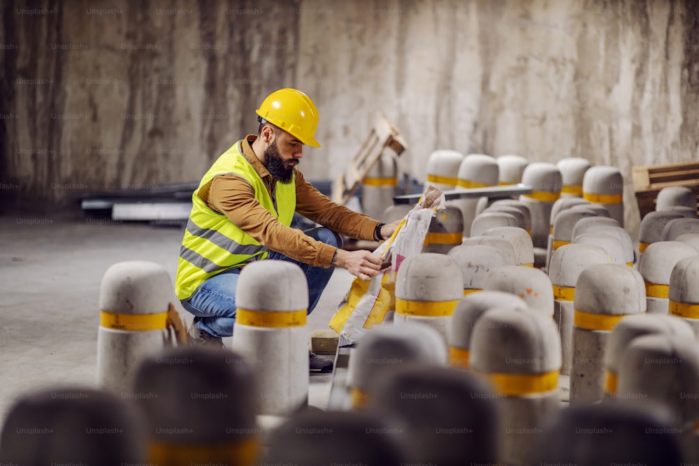 Young attractive bearded worker in working clothes crouching and looking at cement in bag.
