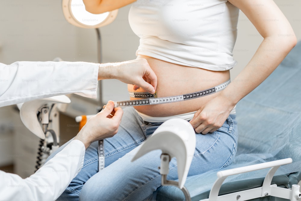 Médecin mesurant le ventre d’une femme enceinte à l’aide d’un ruban adhésif lors d’un examen médical, vue recadrée sans visage focalisé sur l’abdomen