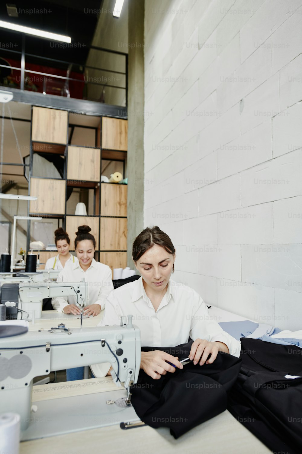 Drei junge Näherinnen sitzen in Reihe an elektrischen Nähmaschinen und arbeiten gemeinsam an neuen Modekollektionen oder Kundenaufträgen
