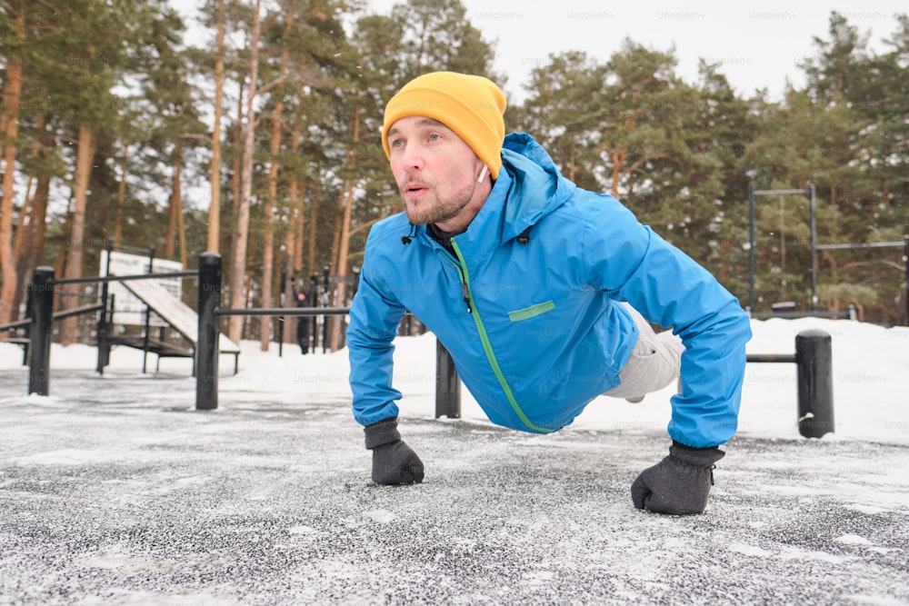 Starker junger Mann mit Ohrstöpseln, der sich beim Training im Trainingsbereich im Winter vom Boden wegdrückt