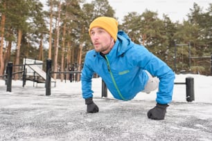 Strong young man in ear buds pushing himself away from ground while training at workout area in winter