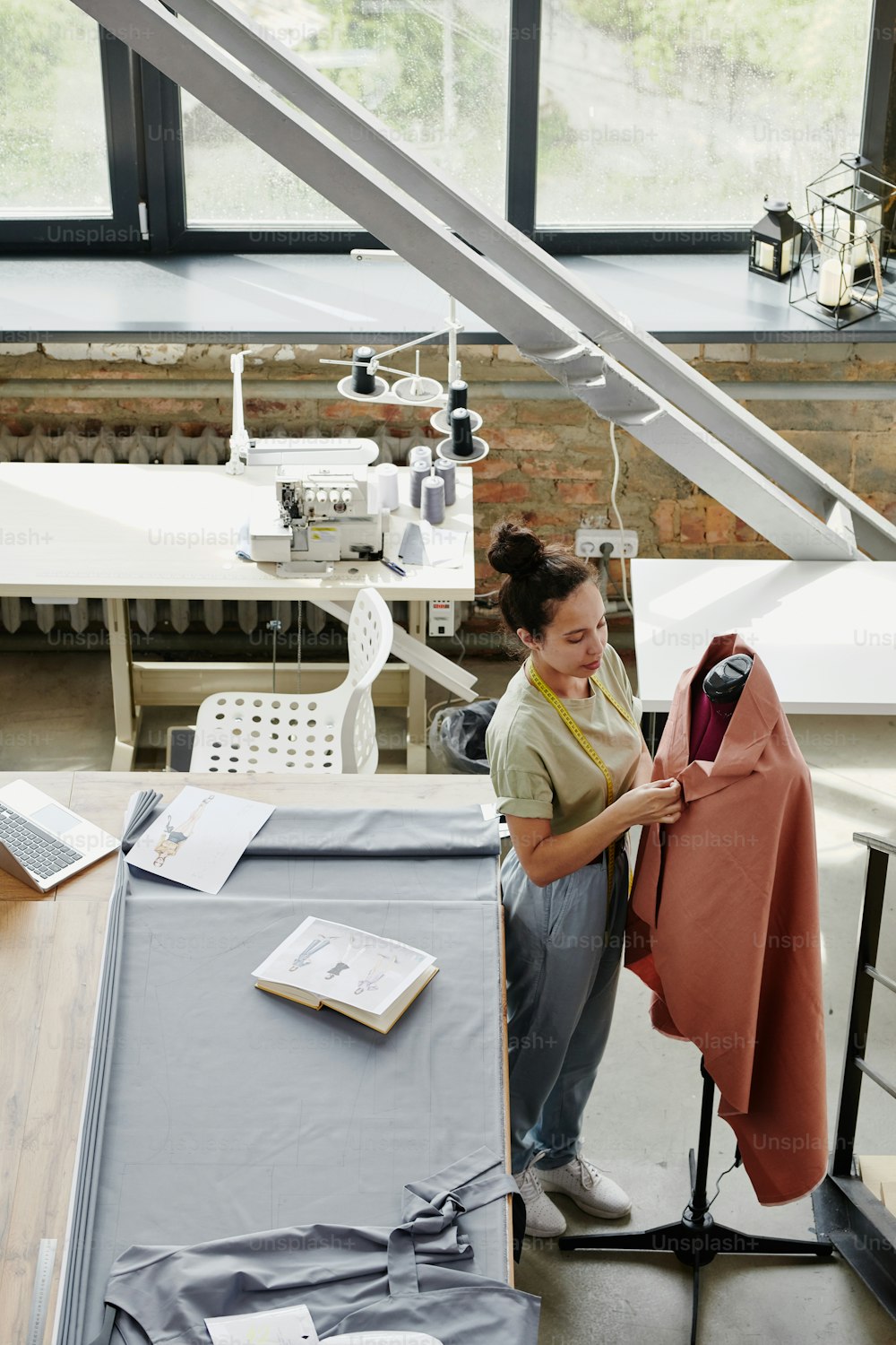 Young contemporary fashion designer working with textile on dummy while standing by table with fabric, open notebook and sketch