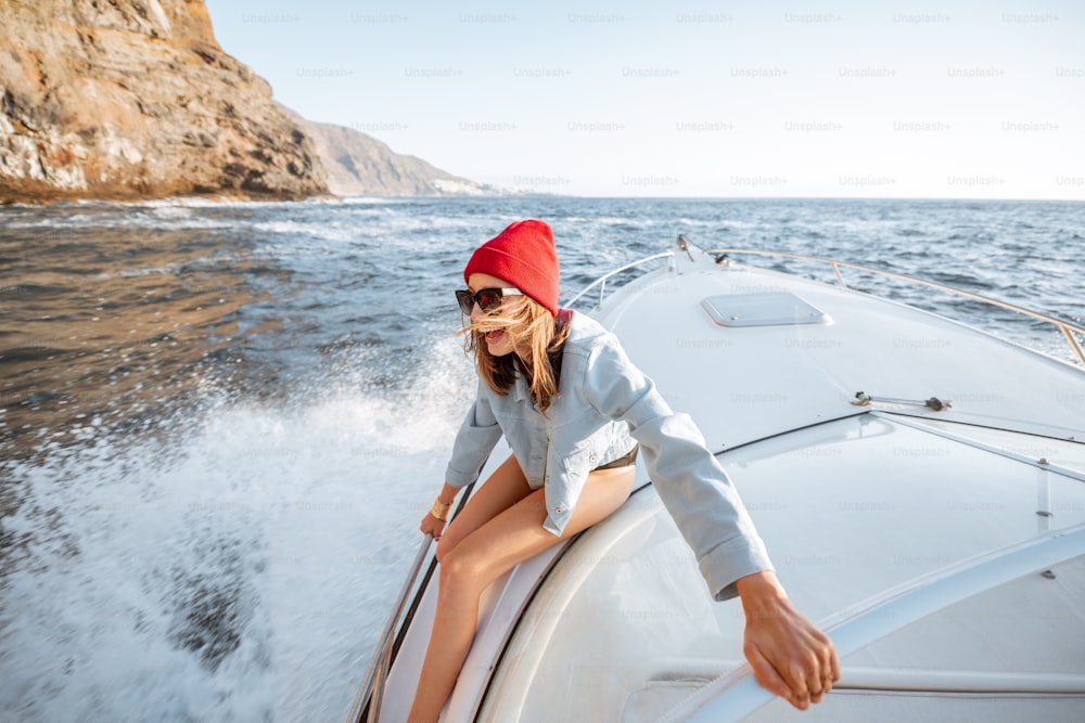 Young woman dressed casually in jacket and red hat enjoying sea voyage, sailing on a yacht in stormy ocean near a rocky shore. Extreme summer recreational pursuit