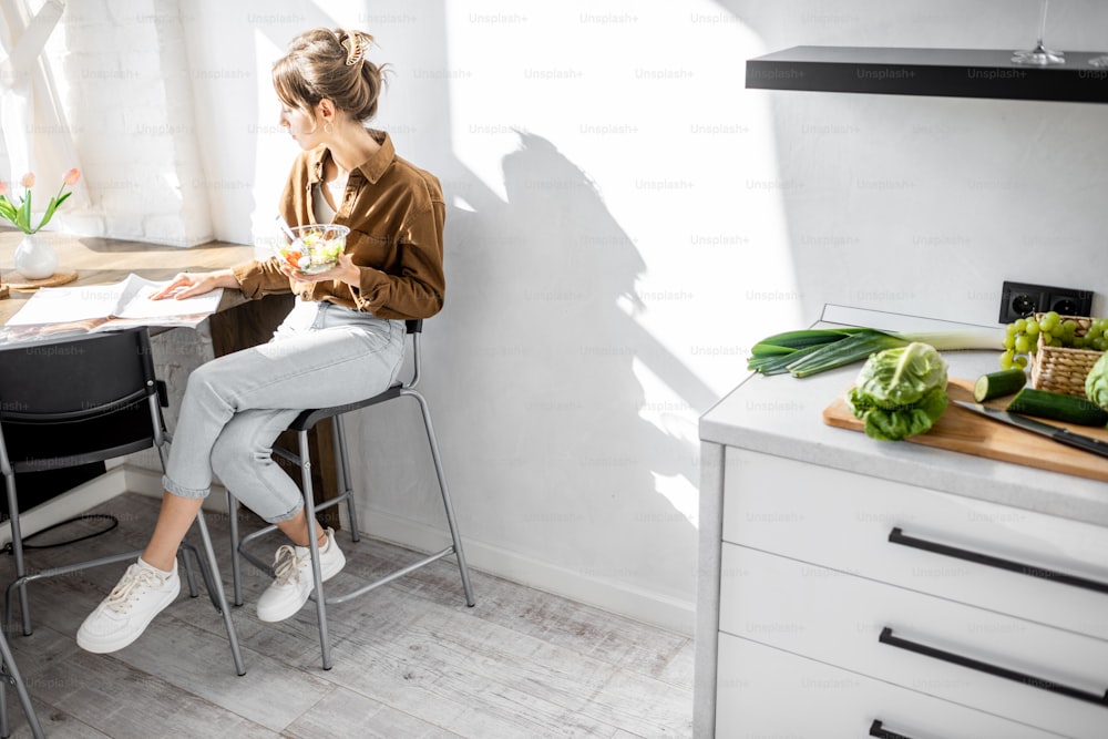 Young woman having a snack with healthy salad, sitting in the white and sunny kitchen at home