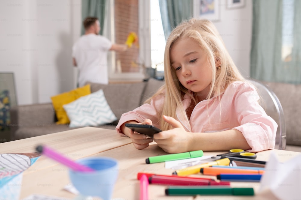 Little restful blond girl scrolling in smartphone while sitting by table in living-room with her father cleaning windows on background