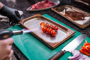 Close-up of unrecognizable chef heating cherry tomatoes on metal tray using gas spray gun while cooking dish