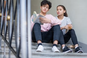 Estudiante inteligente en ropa casual tomando notas en cuaderno mientras está sentado en la escalera con su compañero de clase cerca antes de la lección