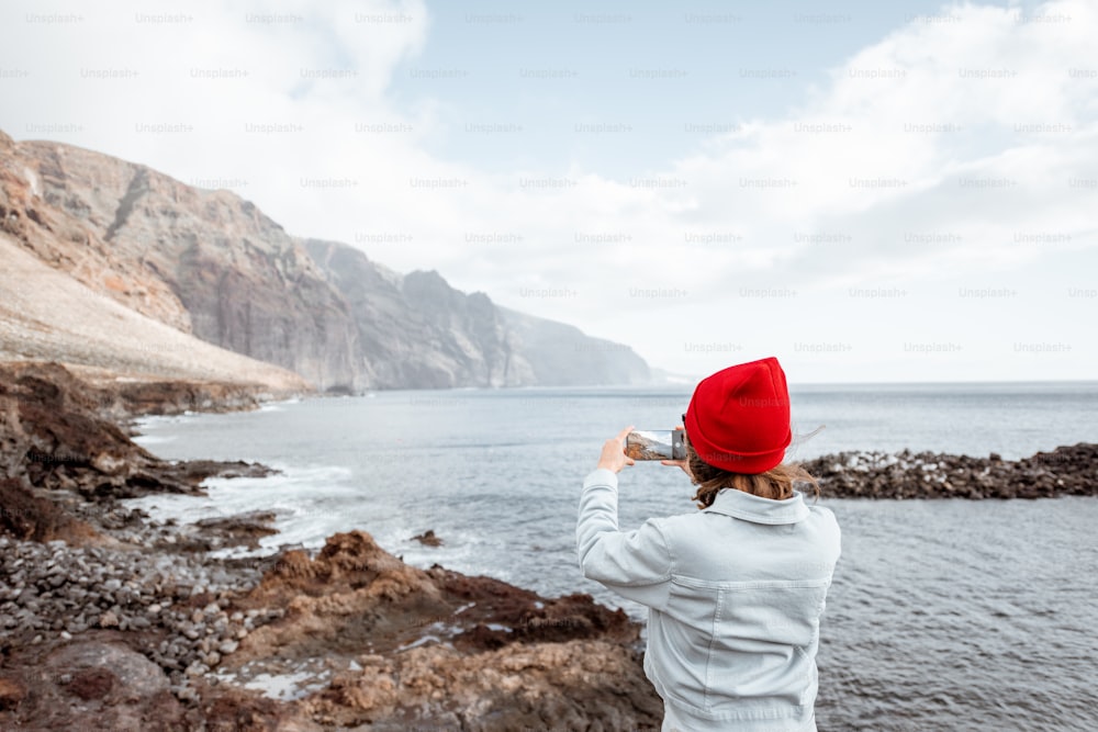 Young woman in red hat enjoying a trip on a rocky ocean coast, photographing on phone breathtaking landscapes. Traveling on Tenerife island, Spain