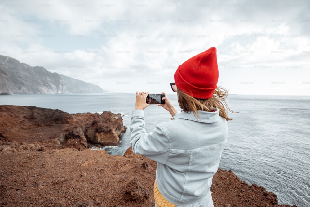 Mujer joven con sombrero rojo disfrutando de un viaje en una costa rocosa del océano, fotografiando por teléfono paisajes impresionantes. Viajar a la isla de Tenerife, España