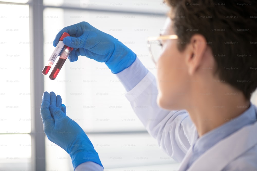 Over shoulder view of female lab specialist in gloves holding test tubes against light while researching blood by coronavirus patient