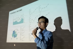 Confident young Asian man in eyeglasses standing at projection screen and speaking into microphone while presenting his business idea to investors