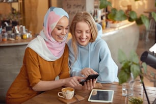 Friends. Young Girls In Cafe. Smiling Women Looking At Smartphone Screen. Different Ethnicity Female Spending Time In Bistro.