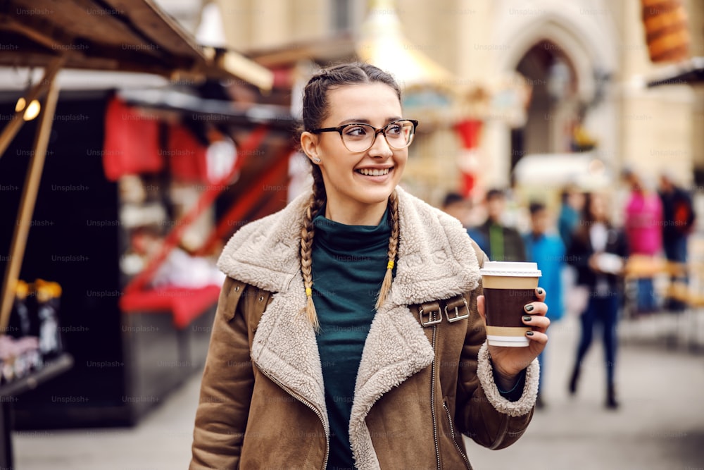 젊은 쾌활한 소녀가 시내 거리를 걷고, 신선한 커피와 함께 일회용 컵을 들고 주말에 자유 시간을 즐기고 있습니다.