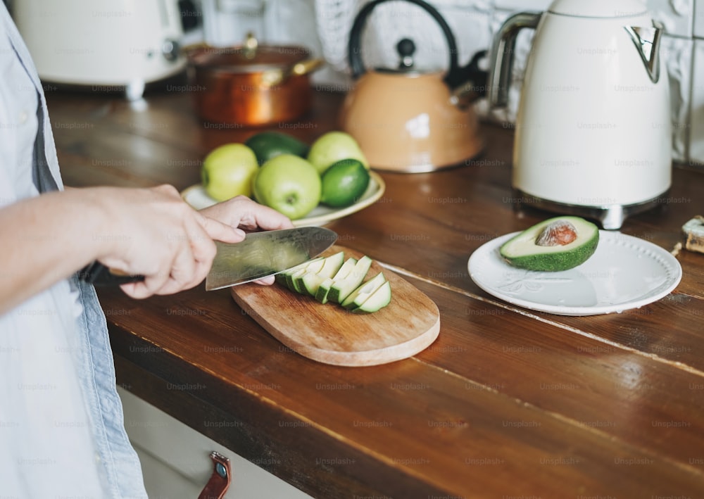 Las manos femeninas cortan aguacate en una superficie de trabajo de madera en la cocina en casa.