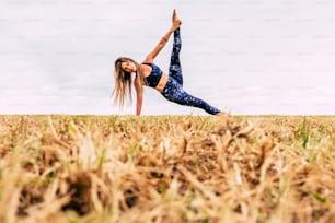 Young beautiful caucasian woman doing balanced pilates fitness position in the outdoor meadow nature - healthy lifestyle and sport activity people concept - white sky background