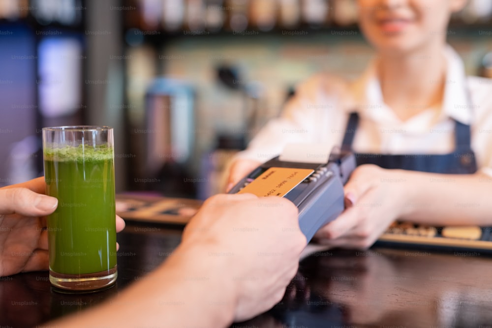 Contemporary client holding plastic card over screen of payment terminal while paying for glass of vegetable smoothie in cafe