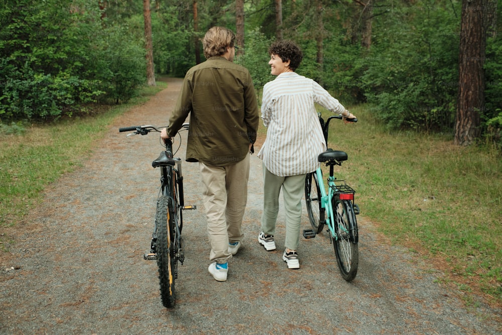 Rear view of happy young husband and wife in casualwear talking to each other while moving down forest path or road after riding bicycles