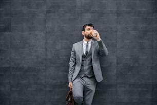 Young attractive bearded fashionable businessman leaning on the wall and drinking his morning coffee.