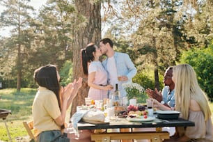 Young amorous couple kissing under pine tree by served table while their friends congratulating them with engagement by clapping hands